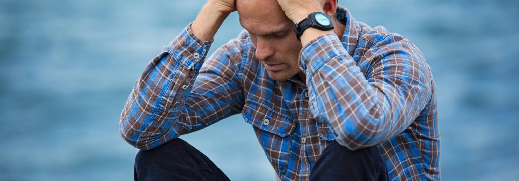 A man in a plaid shirt sits by the water looking distressed, symbolizing stress.